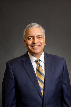 Ajay Vinzé is wearing a dark blue sportcoat and a gold tie with blue diagonal stripes.